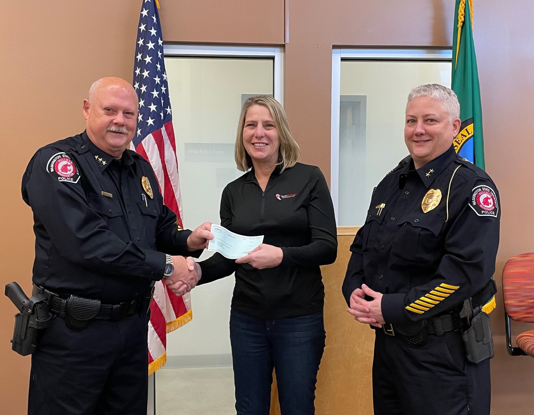 Two Washington State University police officers, Gary Jenkins and Dawn Daniels, stand on either side of Jill McCluskey who is shaking hands with Gary Jenkins. The group is standing in front of an American flag and a Washington State flag.