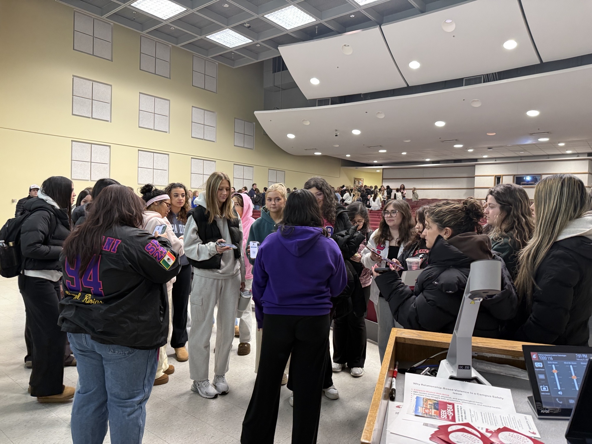 Group of Washington State University Greek life students standing together in a campus lecture hall after a presentation, talking in small groups and holding phones and materials related to a campus safety discussion on relationship violence and stalking.